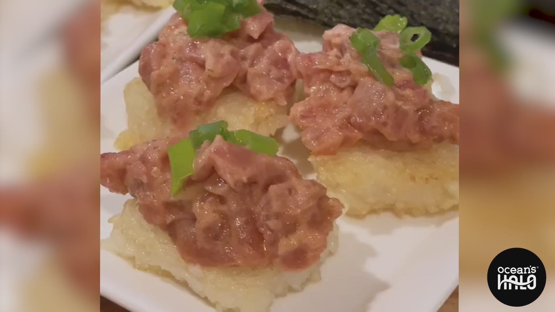 Close-up of sushi featuring three rice squares topped with pink tuna, garnished with green onions, on a white plate.
